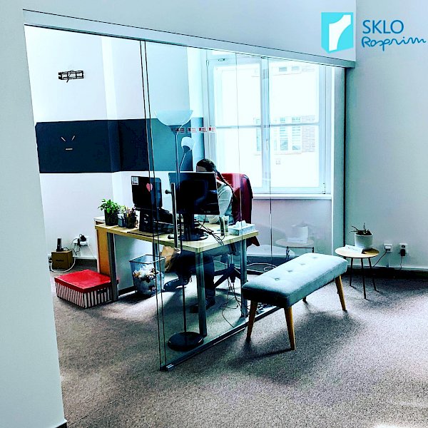 Skleněná zástěna mezi pracovnou a obývákem, průhled do pracovny s pracovním stolem a monitorem, žena sedící na židli za stolem